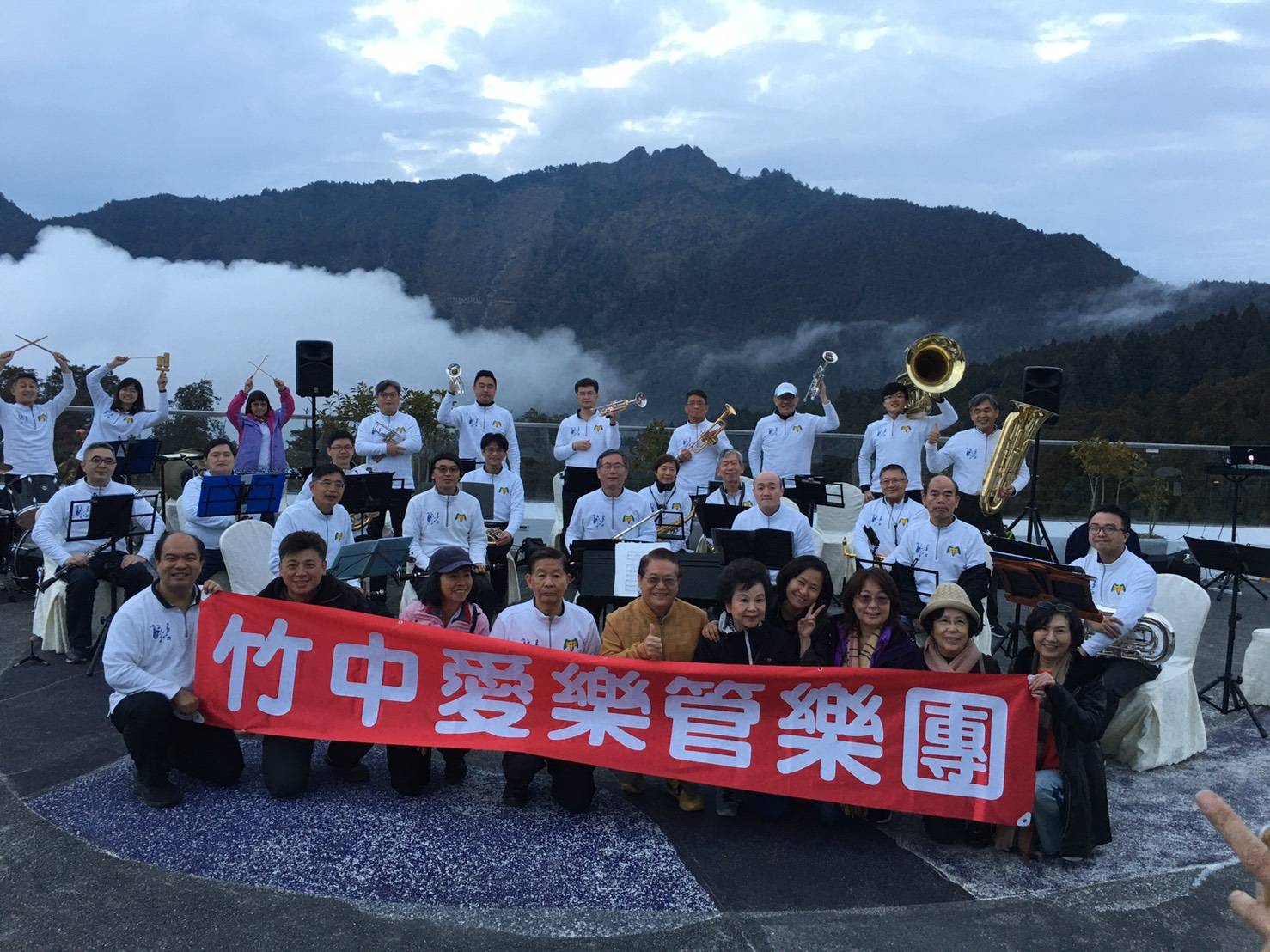 竹中愛樂管樂團創團首次遠征阿里山演出的全團大合照,團員穿著白色團服、拉著紅色團名布條,背景是雲霧繚繞的山景